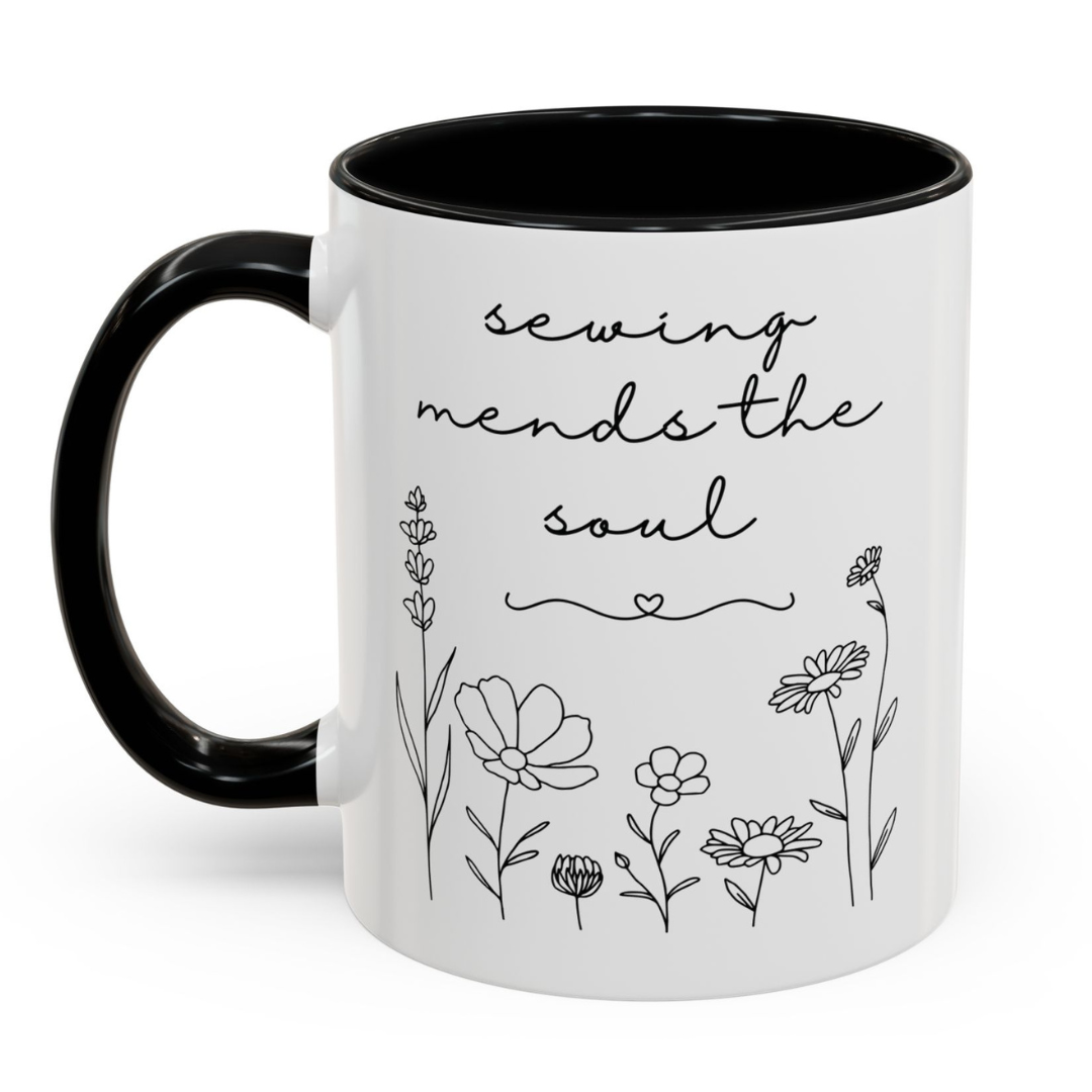 White mug with black interior and handle, featuring floral designs and the text 'sewing mends the soul' on a white background.