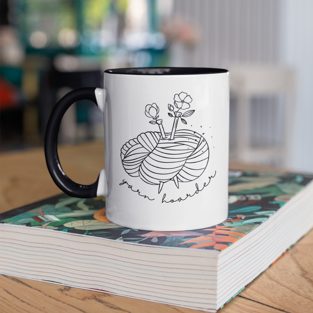 White mug with black handle featuring a knitting design and text, placed on a book.
