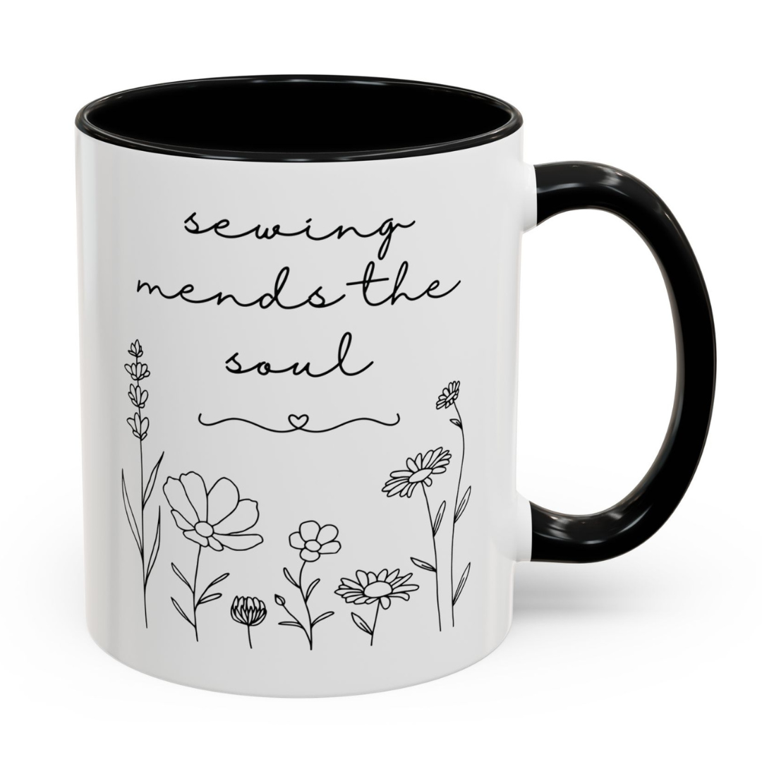 White mug with black interior and handle, featuring floral designs and the text 'sewing mends the soul' on a white background.